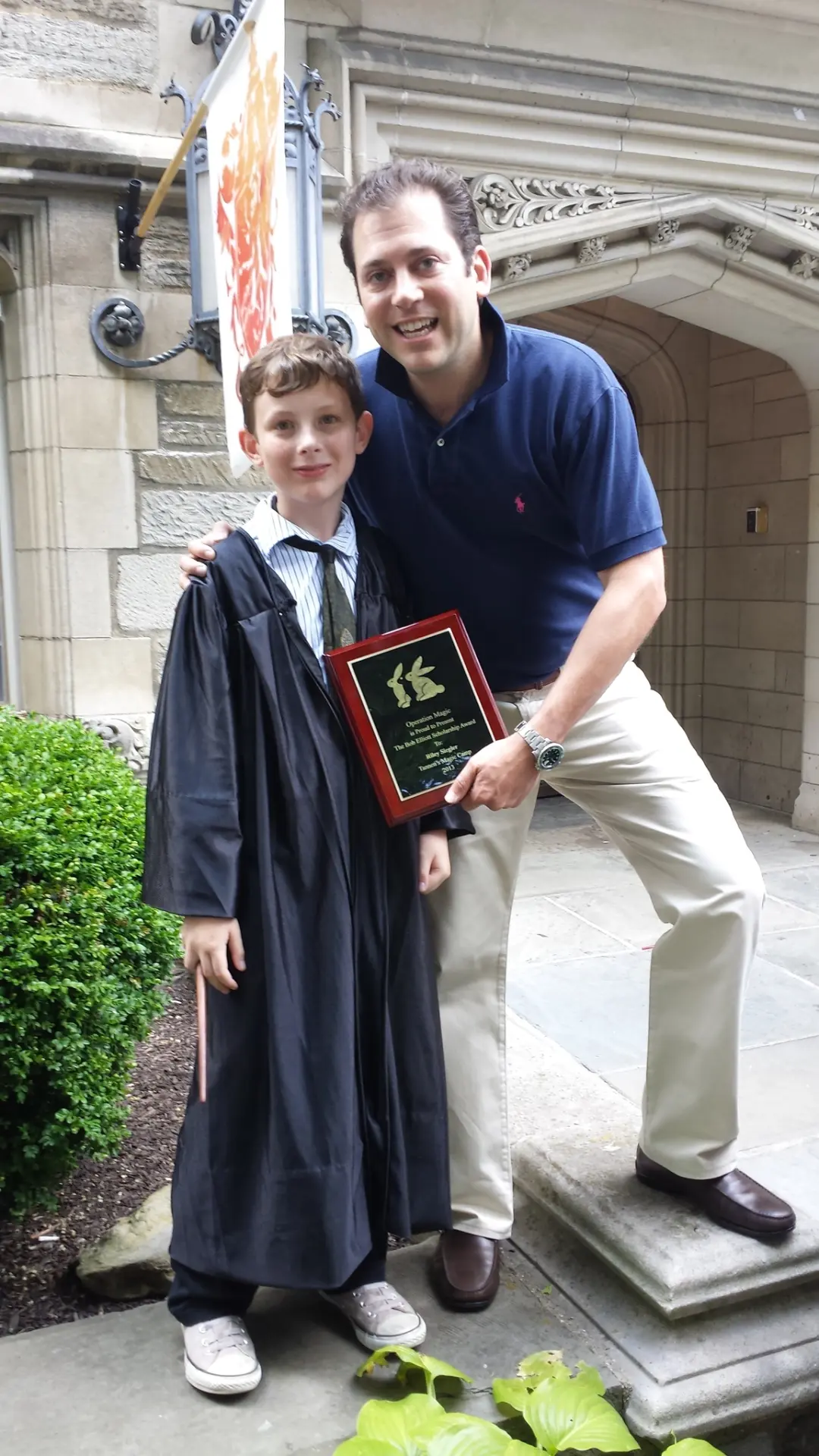 A young magician in a graduation robe holding his Operation Magic scholarship plaque beside a smiling presenter
